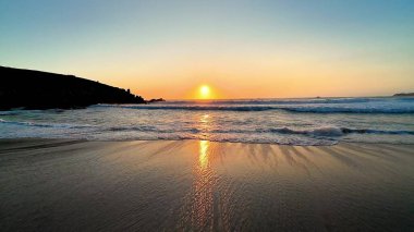 Gün batımının nefes kesici görüntüsü sakin ve durgun sulara güzel bir şekilde yansıyor, sakin bir sahil manzarası yaratıyor.