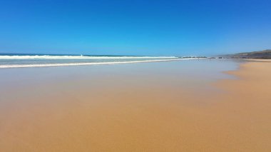 Huzurlu ve sakin bir sahil sahnesi yumuşak altın kum, nazik dalgalar ve açık mavi bir gökyüzü sergiliyor.