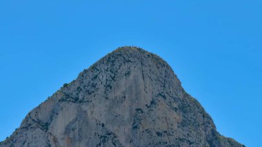 Kayalık bir dağın nefes kesici manzarası yukarıdaki parlak ve berrak mavi gökyüzüne karşı güzel bir şekilde yükseliyor.