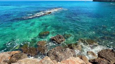 Berrak mavi gökyüzü altında canlı turkuaz suların ve kayalık oluşumların yer aldığı çarpıcı bir okyanus manzarası..