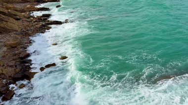 Bu hava manzarası, kayalık kıyı şeridi ve nazik dalgalarla karşılaşan çarpıcı turkuaz suları yakalar.