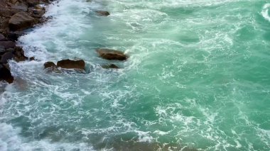 Turkuaz dalgaların büyüleyici ve nefes kesici görüntüsünü deneyimleyin. Sahildeki engebeli taşların üzerinden nazikçe yıkayın.