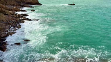 Güzel ve sakin bir sahil manzarası turkuaz suları sergiliyor üzerinde bulutlu bir gökyüzünün altında kayalık uçurumlar var.