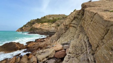 Nefes kesici manzara, canlı turkuaz deniz kıyısında engebeli kayalara çarpan dramatik uçurumları ve dalgaları gösteriyor.