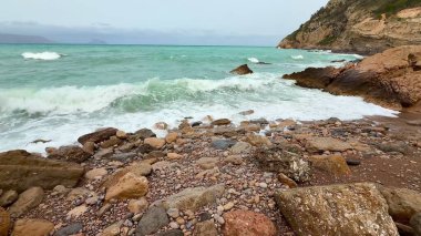 Bulutlu bir gökyüzünün altında engebeli bir kayaya çarpan güçlü dalgaların nefes kesici ve çarpıcı görüntüsü.
