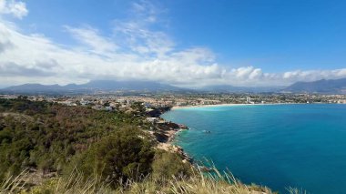 Çarpıcı bir kıyı manzarası parlak, güneşli bir gökyüzünün altındaki görkemli dağların yanında berrak mavi suları gösteriyor.