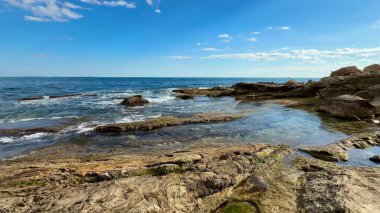 Berrak mavi suları, engebeli kayaları ve güzel boyanmış gökyüzünü gösteren sakin bir sahil manzarası.