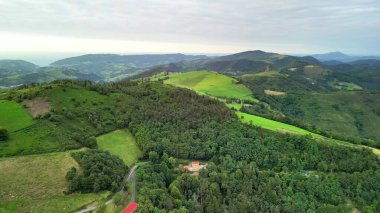 Hava manzarası, bulutlu bulutlu gökyüzünün altında yemyeşil tepeleri ve altın tarlaları yakalıyor ve sakin bir manzara yaratıyor.