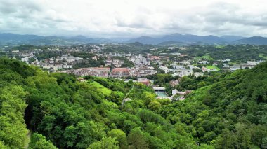 Büyüleyici bulutlu gökyüzünün altında yemyeşil tepelere yuva yapmış canlı bir şehrin çarpıcı hava manzarası.