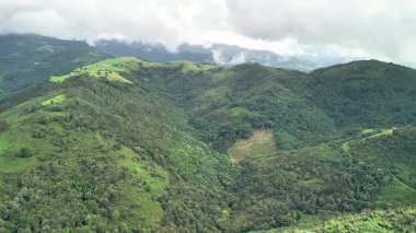Hava manzarası, dramatik bulutlu bir gökyüzünün altında yemyeşil dağlar ve vadileri gösteriyor. Resimli bir manzara yaratıyor.