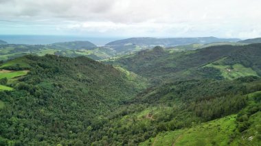 Uçsuz bucaksız okyanus manzaralarının çerçevelediği yemyeşil vadileri, bulutlu bulutlu gökyüzünün altında bir hava manzarası vurgular.