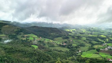 Göz kamaştırıcı bir hava manzarası yemyeşil manzaraları ve geniş bulutlu gökyüzünün altında yuvarlanan tepeleri gösteriyor.