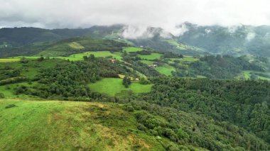 Hava manzarası canlı yeşillikler, yuvarlanan tepeler ve bulutlu bir gökyüzünün altında görkemli dağlar sergiliyor.