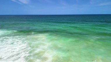 Berrak mavi gökyüzünün altında sersemletici turkuaz suları ve nazik dalgaları gösteren nefes kesici bir deniz manzarası.