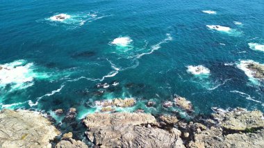 Okyanus dalgalarının canlı mavi gökyüzünün altında kayalık kıyılara çarptığı çarpıcı hava görüntülerini tecrübe edin.