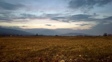 Sakin bir alacakaranlık manzarası engin tarlaları ve uzak tepeleri sergiliyor bulutlu bir akşam gökyüzünün altında zarafetle.