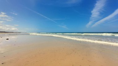 Yumuşak kum, yumuşak dalgalar, parlak mavi bir gökyüzünün yanında hoş ve huzurlu bir sahil sahnesi.