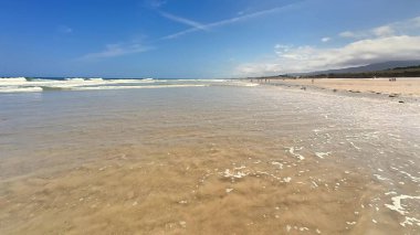 Bu büyüleyici bir plaj sahnesi kristal berrak sular, yumuşak altın kum ve parlak mavi gökyüzü