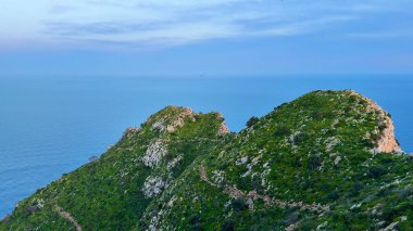 Bereketli yeşil tepelerden oluşan nefes kesici bir manzara. Pastel gökyüzünün altında sakin okyanus sularıyla güzel bir şekilde buluşuyor.