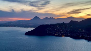 Gün batımında nefes kesici bir kıyı kenti manzarası, görkemli dağlar ve suya yansıyan parlak ışıklar.