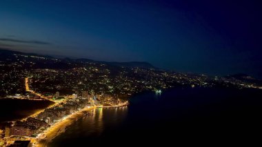 Nefes kesici bir hava manzarası canlı şehir ışıklarını ve alacakaranlıkta kıyı şeridini gözler önüne seriyor, kentsel güzelliği vurguluyor..