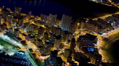 Gece canlı bir kıyı şehrinin büyüleyici hava manzarası aydınlanmış binalar ve sakin sular sergileniyor..