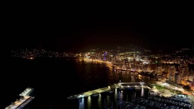 Canlı bir sahil şehrinin nefes kesen manzarası gece aydınlanıyor, şehrin ışıklarını ve liman manzarasını sergiliyor..