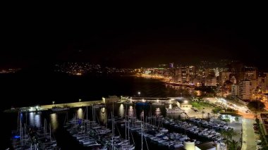 Göz kamaştırıcı bir gece limanı sahnesi aydınlık yatları ve körfez boyunca ışıl ışıl parlayan şehir silüeti.