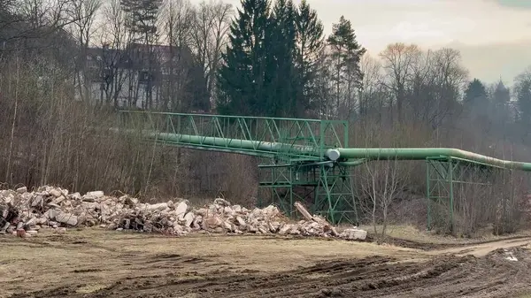 Endüstriyel alan boyunca uzanan uzun yeşil bir konveyör köprüsü ön planda çeşitli enkaz parçalarıyla birlikte...