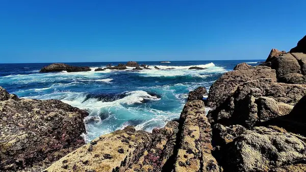 Nefes kesen okyanus manzarası, açık mavi gökyüzünün altında kayalara çarpan muhteşem dalgaları sergiliyor.