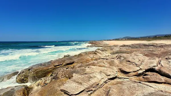 Büyüleyici bir sahil sahnesi görkemli kayalar, yumuşak okyanus dalgaları ve güzel açık mavi bir gökyüzü sergiliyor.