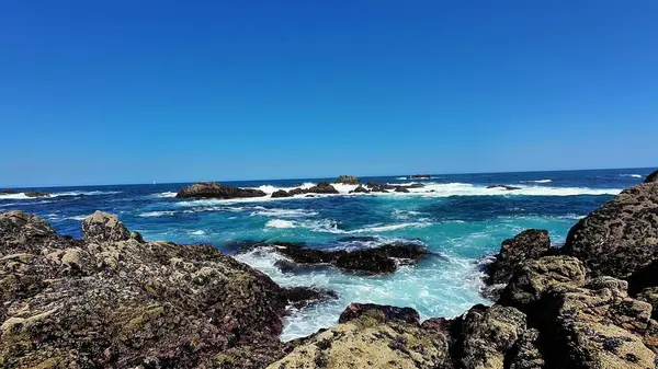 Dalgaların mavi gökyüzünün altında kayalara çarptığı çarpıcı bir sahil manzarası. Plaj keşfi ve rahatlama için mükemmel.