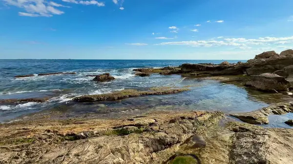 Berrak mavi suları, engebeli kayaları ve güzel boyanmış gökyüzünü gösteren sakin bir sahil manzarası.