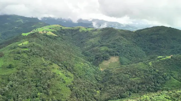 Hava manzarası, dramatik bulutlu bir gökyüzünün altında yemyeşil dağlar ve vadileri gösteriyor. Resimli bir manzara yaratıyor.