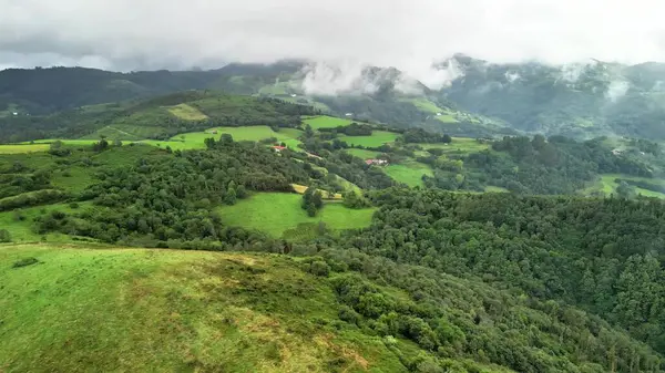 Hava manzarası canlı yeşillikler, yuvarlanan tepeler ve bulutlu bir gökyüzünün altında görkemli dağlar sergiliyor.