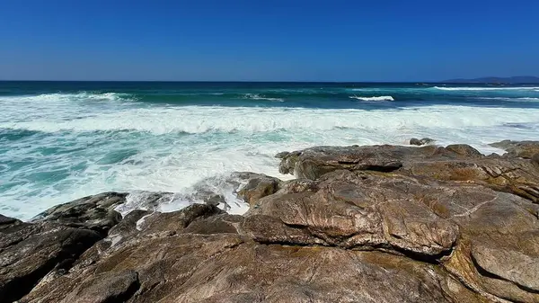 Parlak mavi gökyüzünün altında engebeli bir kıyı şeridine çarpan canlı okyanus dalgalarının nefes kesici görüntüsü.