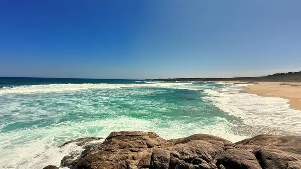 Canlı mavi bir gökyüzünün altında yumuşak dalgalar ve kristal berraklığında sular sergilenen nefes kesici bir sahil manzarası..