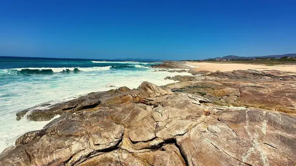 Resimli bir plaj sahnesi engebeli kayalık oluşumlar sergiliyor ve parlak mavi gökyüzünün altında dalgalar çarpışıyor.