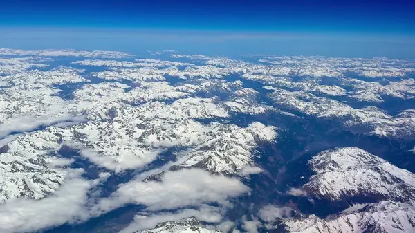 Bu çarpıcı hava görüntüsü, açık mavi gökyüzünün altındaki görkemli karla kaplı dağ zirvelerini güzel bir şekilde vurguluyor.