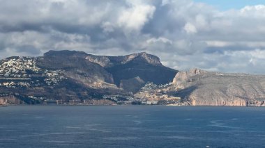 Nefes kesici bir manzara, dramatik bulutlu bir gökyüzü altında görkemli dağlar ve çarpıcı kıyı evleri barındırır.