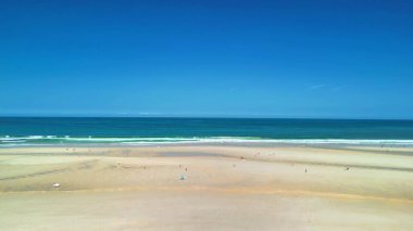 Göz kamaştırıcı bir sahil sahnesinde parlak mavi gökyüzünün altında altın kumlar ve kristal berrak sular sergileniyor.