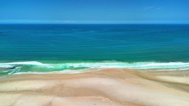 Bu nefes kesici hava görüntüsü, berrak mavi gökyüzünün altında usulca yuvarlanan sakin sahil dalgalarını yakalar.