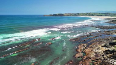 Uçsuz bucaksız okyanus, el değmemiş plaj ve güneşli gökyüzünün altındaki engebeli sahil manzarası.