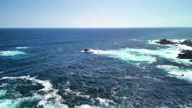 Açık mavi gökyüzünün altındaki kayalık bir sahile çarpan okyanus dalgalarının şaşırtıcı güzellikteki manzarasının tadını çıkar.