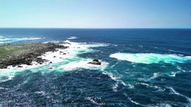 Sakin mavi gökyüzünün altındaki kayalık kıyıların ve canlı turkuaz dalgaların nefes kesici ve çarpıcı hava manzarası.