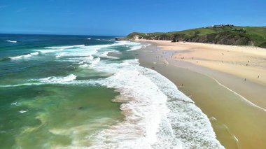 Göz kamaştırıcı bir hava manzarası, açık mavi gökyüzünün altında altın kumları ve canlı dalgaları olan güzel bir plajı sergiliyor.