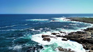 Okyanus dalgalarının berrak mavi gökyüzünün altında kayalık kıyılara çarpışına tanık olun.