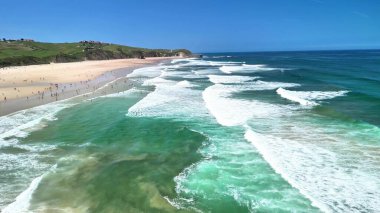 Bu güzel plaj sahnesinde nazik dalgalar, sörfçüler ve güneşlenenler sahilde rahatlatıcı bir günün tadını çıkarıyorlar.