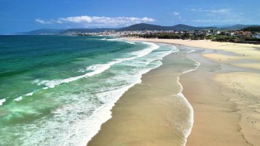 Temiz turkuaz suları ve kıyıya vuran hafif dalgaları olan el değmemiş bir sahilin nefes kesici hava manzarası.