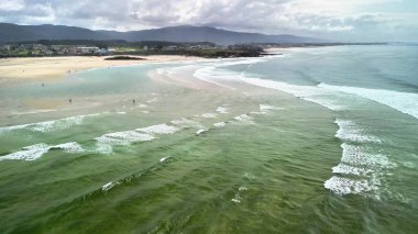 Güzel bir hava manzarası var. Huzurlu bir sahil. Arkasında hafif dalgalar ve dağlar var..
